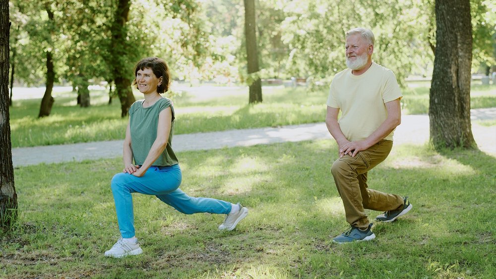 Two people doing lunges together on grass in a park.