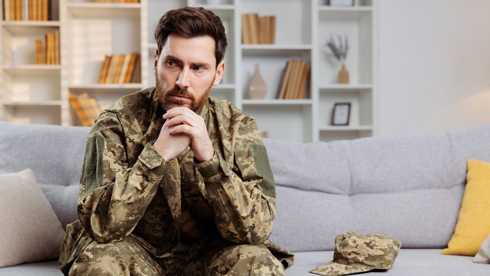 A service member in camouflage sits on a couch with hands clasped, looking down.