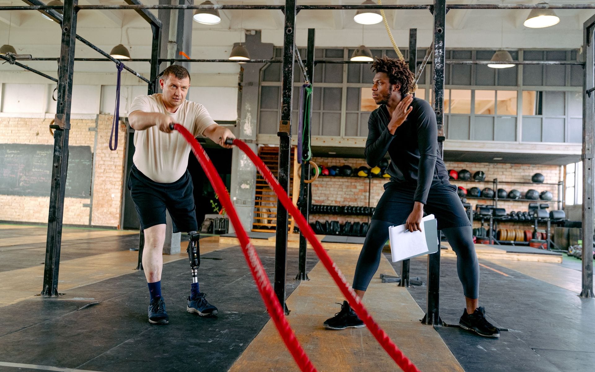 A coach observing an athlete with a prosthetic leg using battle ropes in a gym.