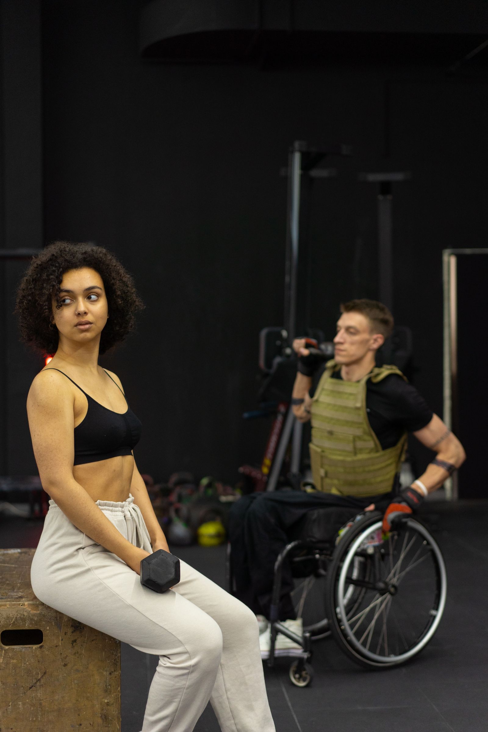 A woman rests on a box holding a dumbbell while a man in a weight vest sits in a wheelchair behind her.