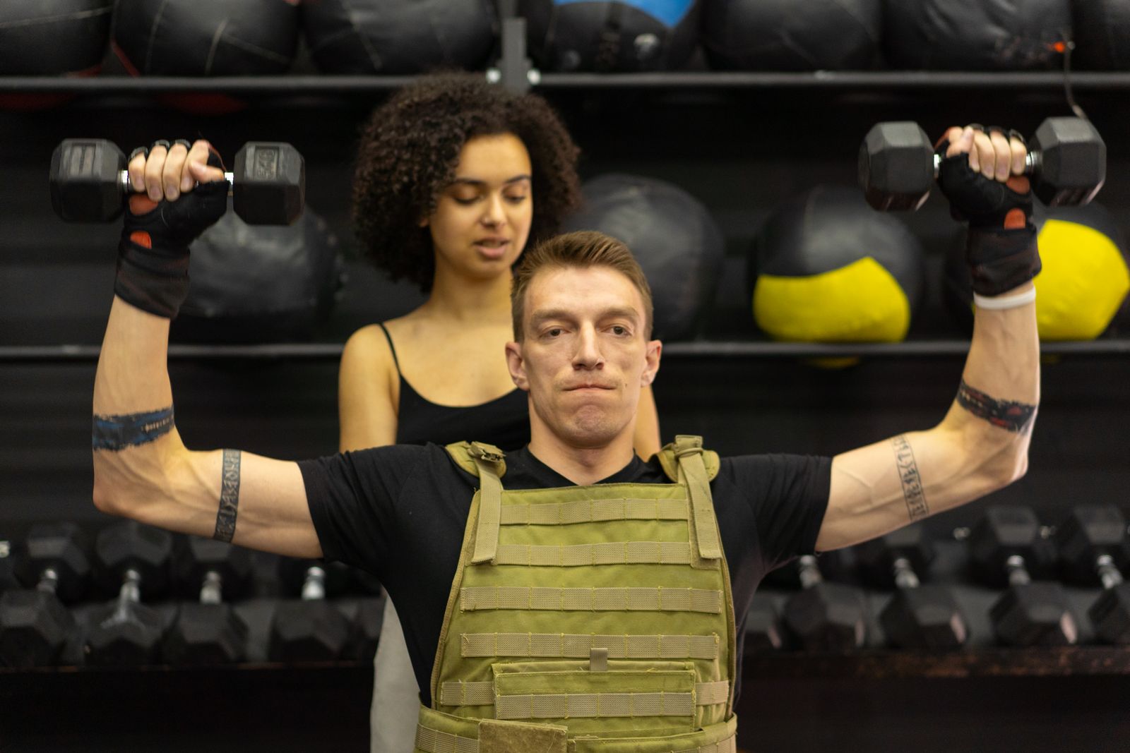 A man in a weight vest presses dumbbells overhead while a coach watches.